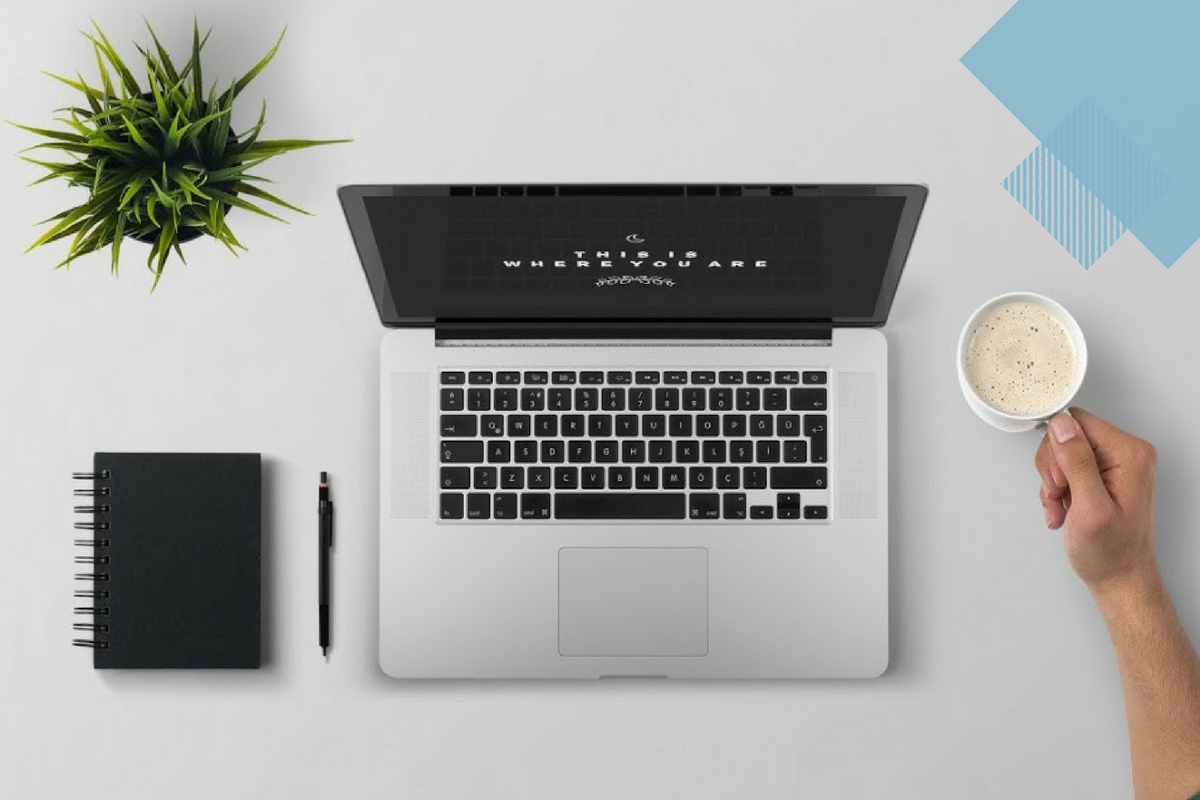 computer sitting on tables with a notebook, pen, and plant next to it. Arm holding a coffee cup is also lying on the table