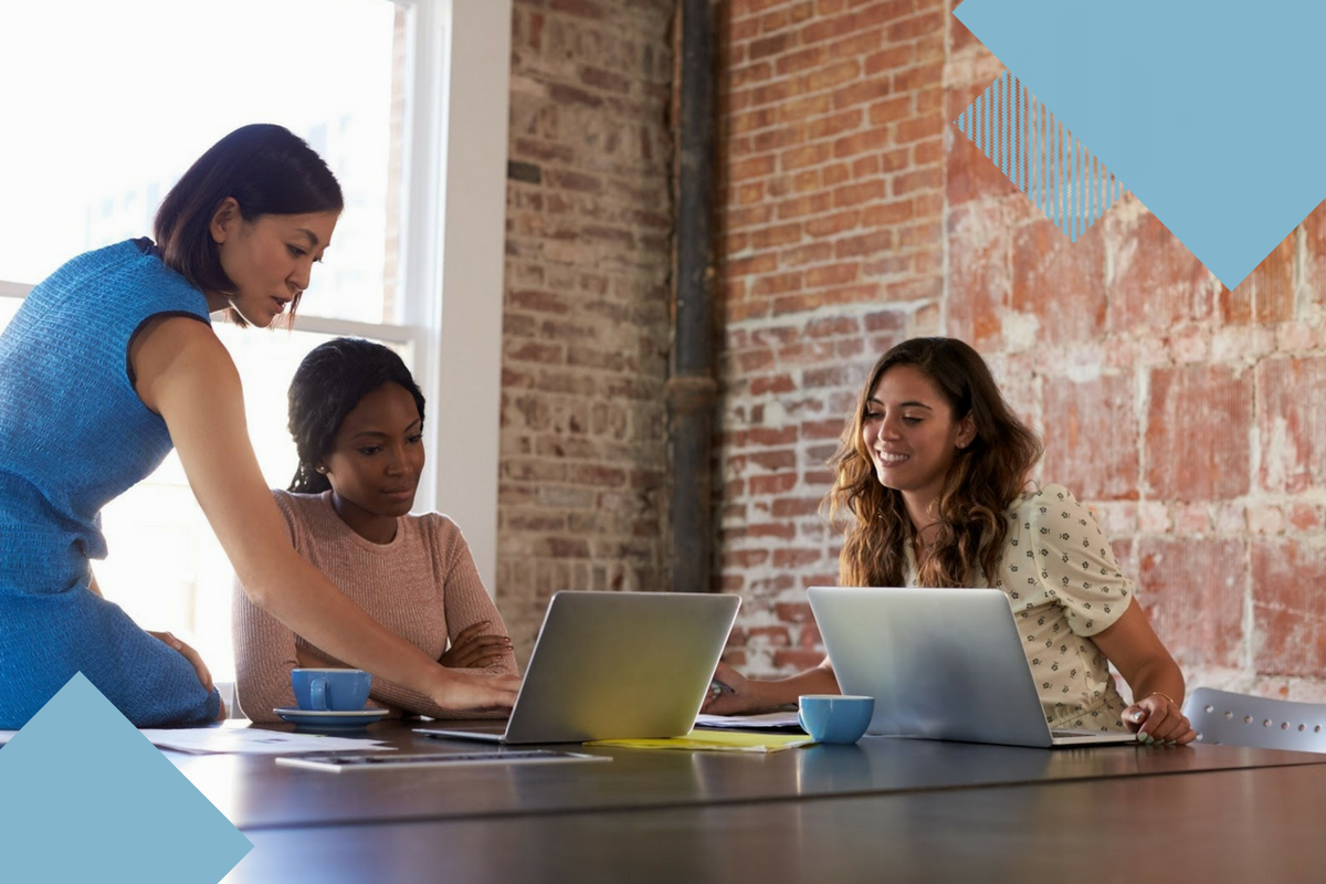 three women working on computers sitting at a large table, with light blue geometric shapes around outside of picture